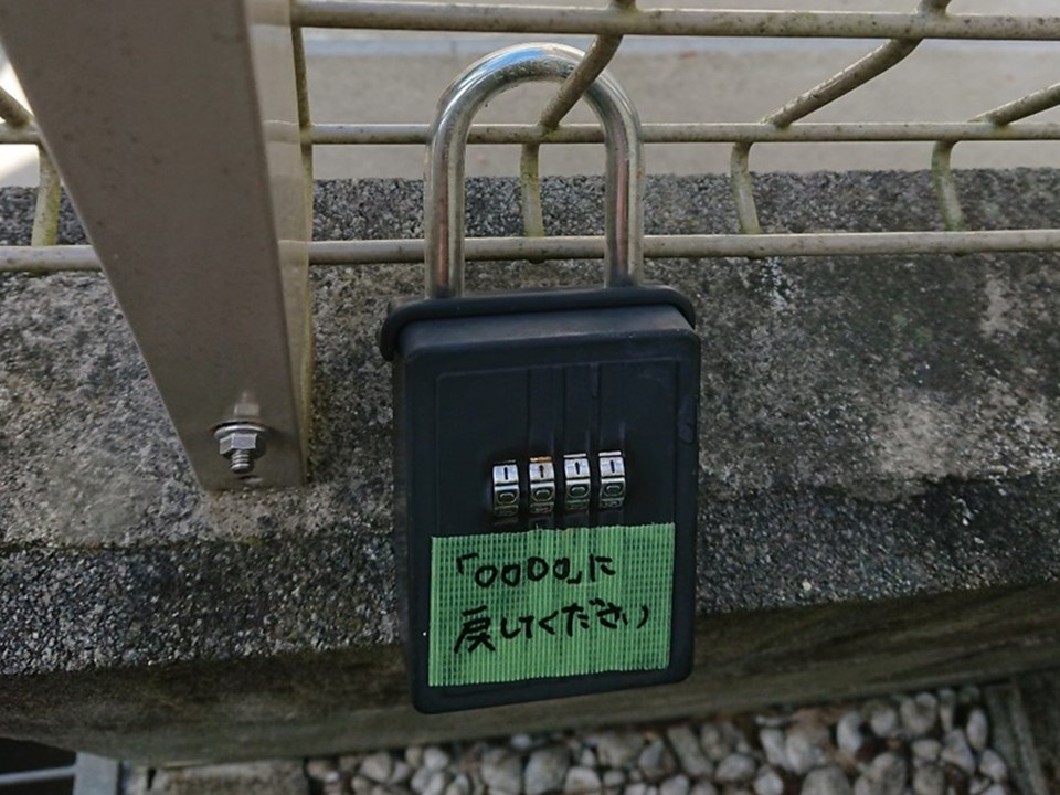 キーボックスの設置例。4桁ダイヤル式のキーボックスをフェンスに取り付け、「0000に戻してください」と書かれた緑色のテープを貼っている