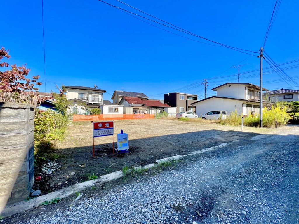 売地の現地設営完了後の全景。敷地手前に「売地」のプレート看板と物件情報ボックス看板を設置し、奥の隣地境界沿いにオレンジ色のネットフェンスを張っている。住宅街の中の整地された更地で、快晴の青空が広がっている