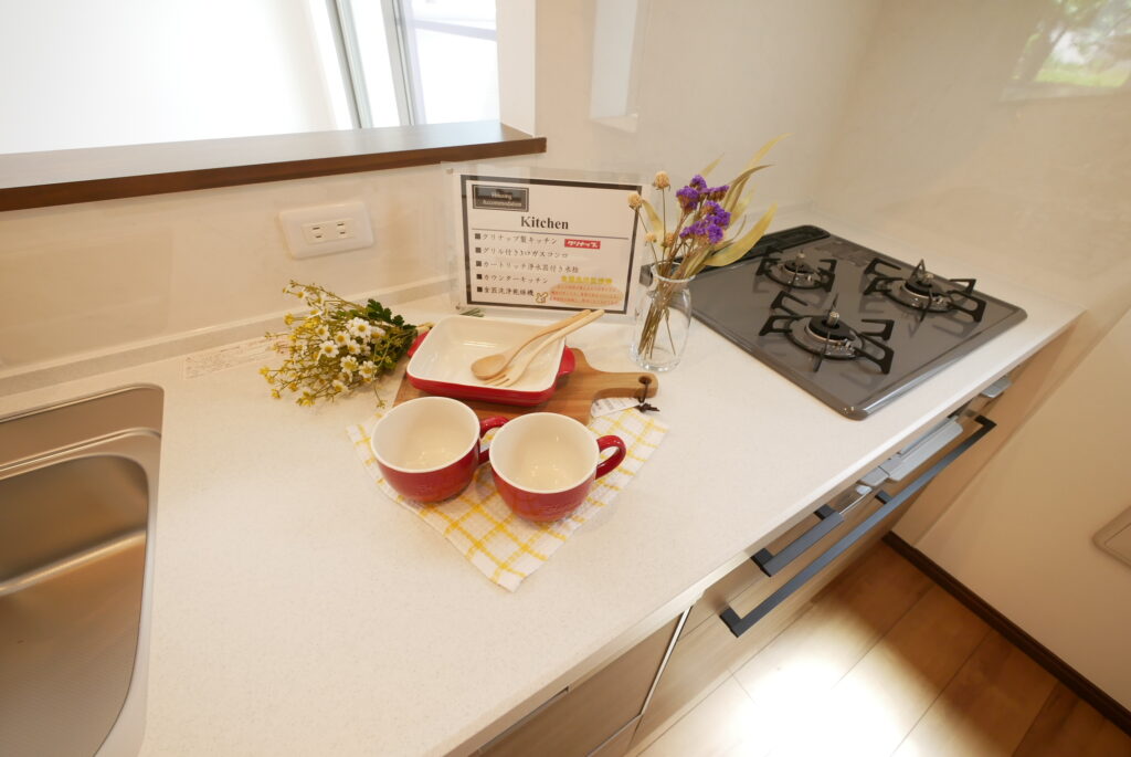 キッチンの室内装飾例。カウンター上に赤いマグカップ2客・木製カッティングボード・赤いグラタン皿・花束・ドライフラワーを配置し、設備仕様を記載したPOPスタンドを設置している