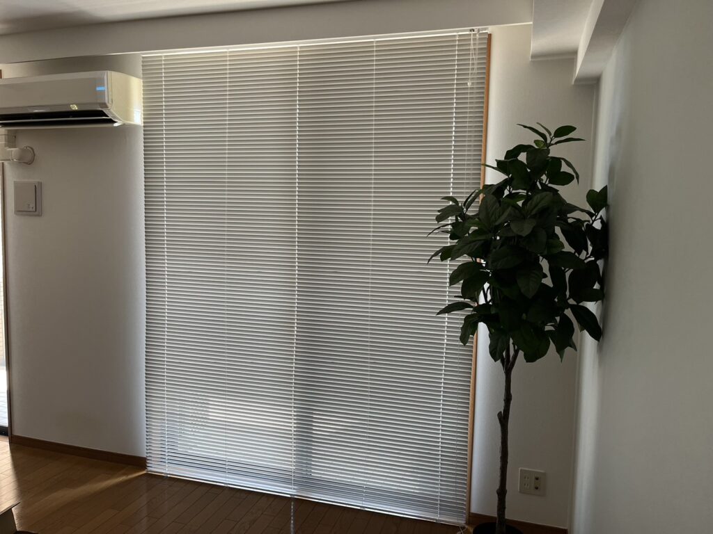 室内装飾のブラインド設置例。リビングの大きな窓にホワイトの横型ブラインドを取り付け、窓辺にフェイクグリーンの大型観葉植物を配置している