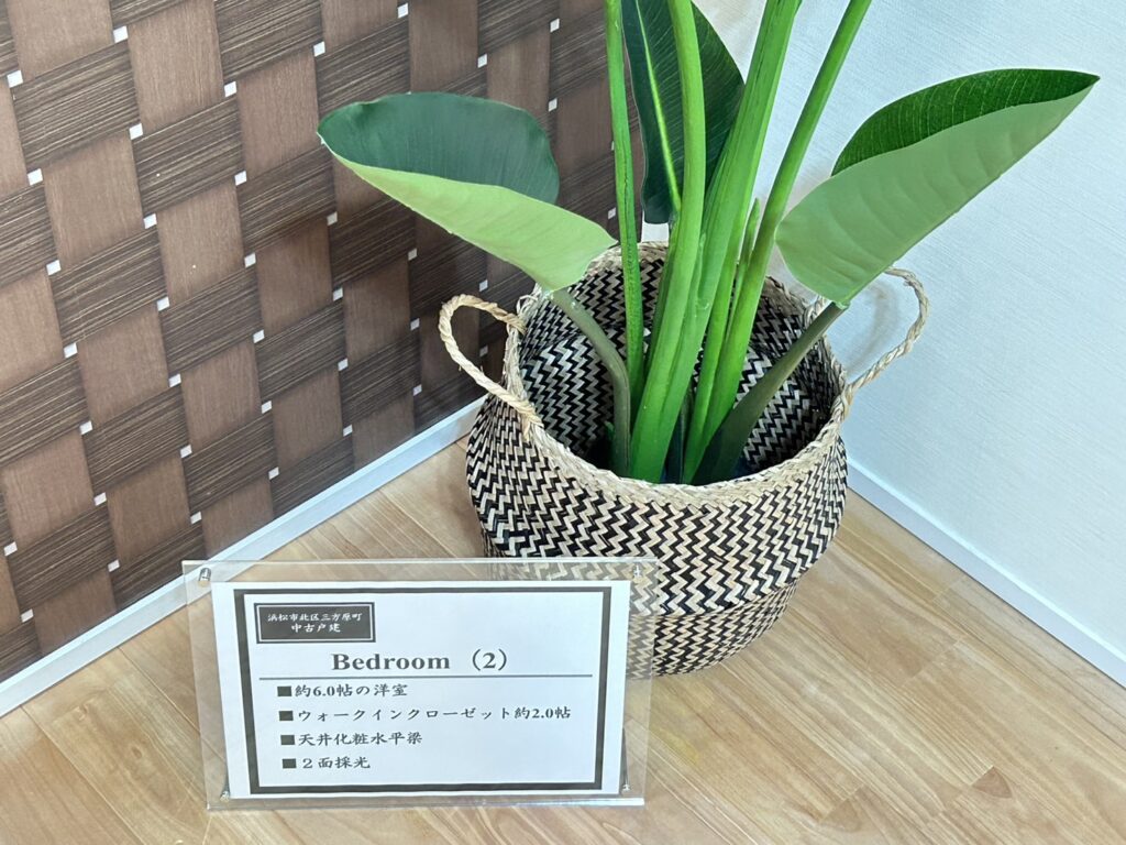 居室コーナーの室内装飾例。ウォールナット調アクセントウォールの前にジグザグ模様のバスケット入り観葉植物を置き、Bedroom（2）の設備仕様を記載したアクリルPOPスタンドを設置している