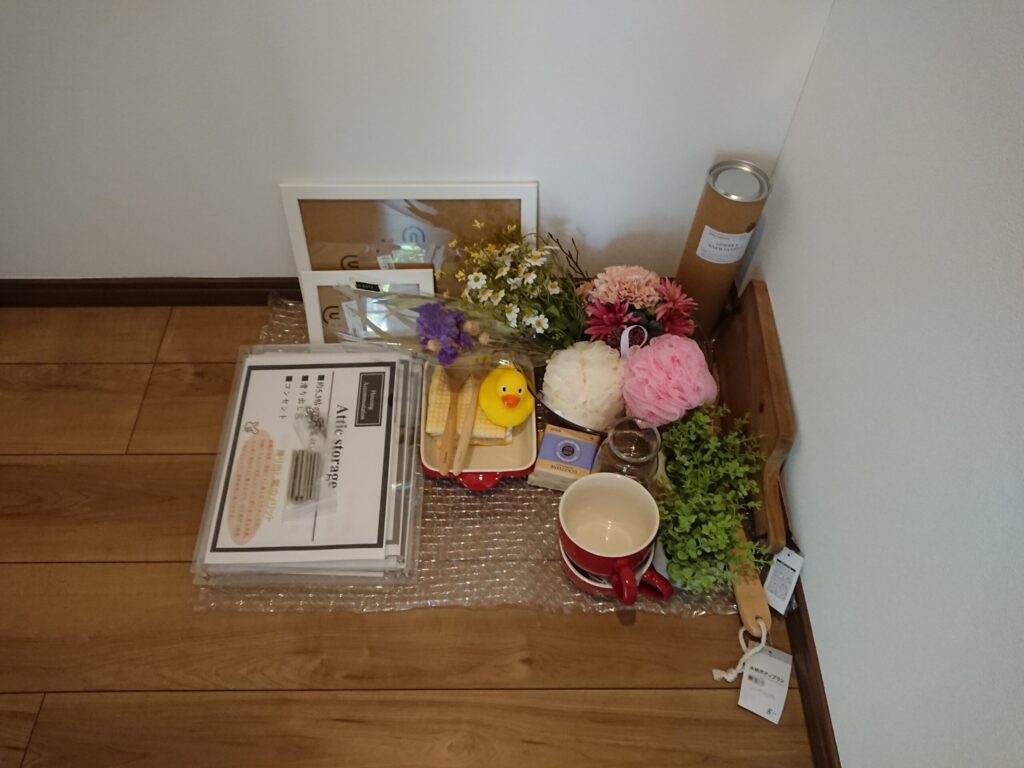 室内装飾用の全アイテム一覧。アクリルPOPスタンド・フォトフレーム・造花・ドライフラワー・フェイクグリーン・赤いマグカップ・赤いグラタン皿・木製スプーン・アヒルのおもちゃ・ボディスポンジ・ボディブラシ・アロマキャンドル缶・カッティングボードなどをまとめて床に並べている