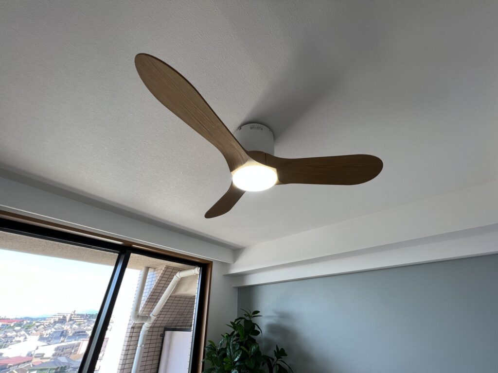 室内装飾のシーリングファン設置例。木目調の3枚羽根シーリングファンライトをリビング天井に取り付け、アクセントクロスの壁と観葉植物で空間を演出している