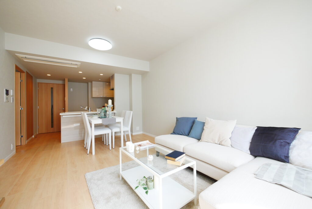 LDKのホームステージング実施例。白いL字ソファにネイビーとベージュのクッションを配置し、ガラス天板のセンターテーブル・白いダイニングテーブルセット4人掛け・ラグを設置して、明るく清潔感のある空間にコーディネートしている