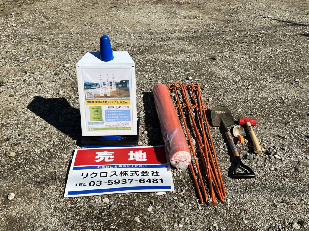 更地の現地設営用アイテム一式。物件情報チラシを入れたボックス看板・「売地」プレート看板・オレンジ色のネットフェンスロール・鉄製ペグ4本・ハンマー・スコップを砂利の地面に並べている
