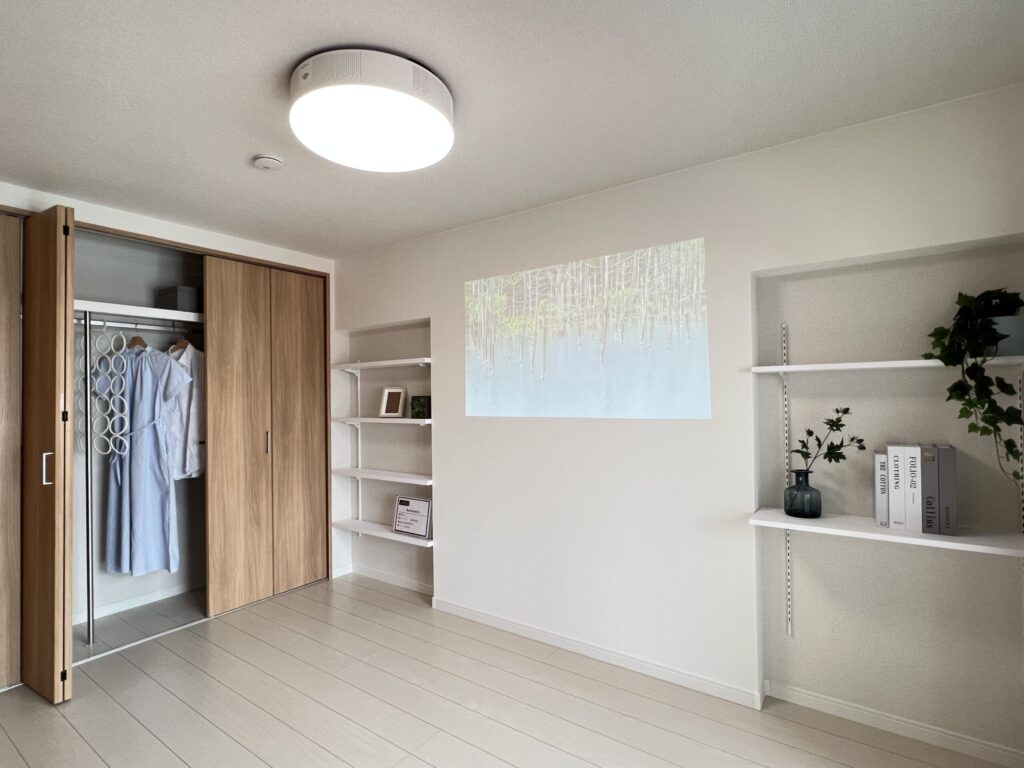 洋室の室内装飾例。木目調のクローゼット扉を開けて衣類をディスプレイし、壁面の棚にアートパネル・花瓶・書籍・観葉植物を配置している