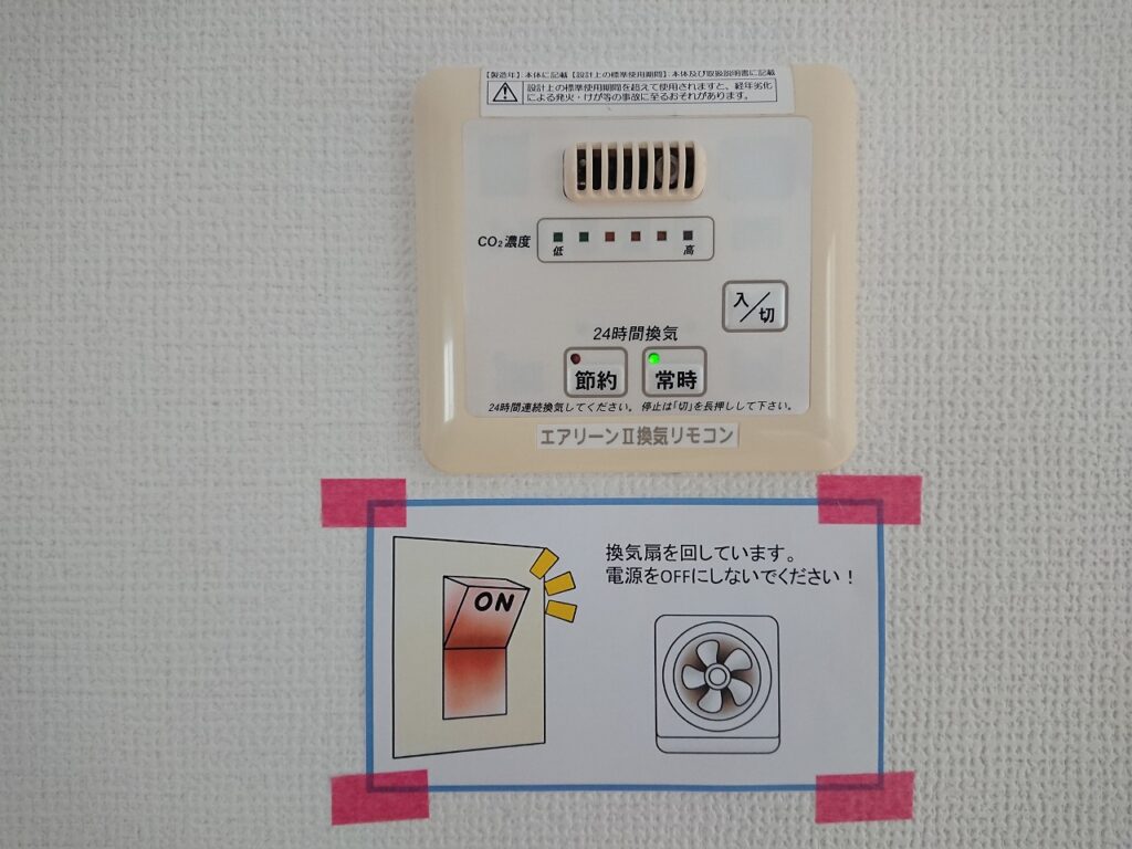 24時間換気リモコンと注意書きの掲示例。エアリーンII換気リモコンの下に「換気扇を回しています。電源をOFFにしないでください！」と記載したイラスト付きの案内を貼り付けている