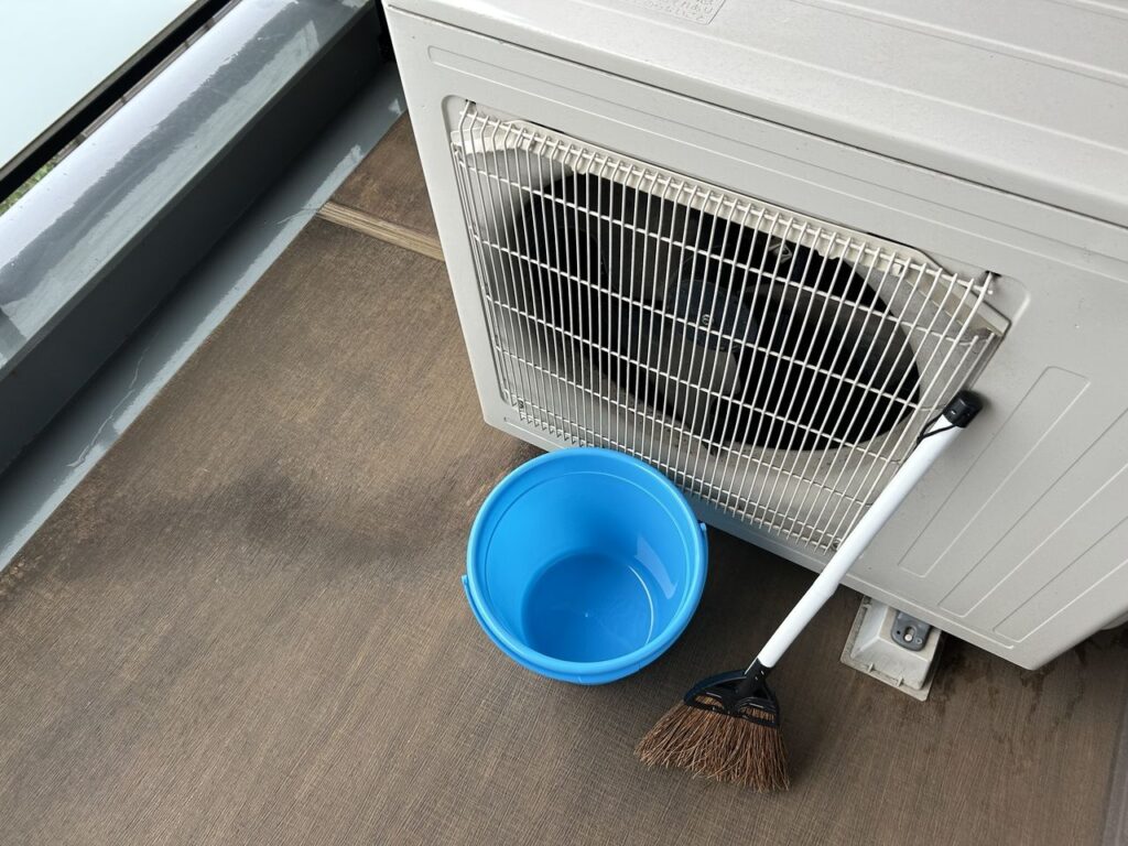 バルコニー清掃の様子。エアコン室外機の横にバケツとほうきを置き、床面の汚れを掃き掃除している