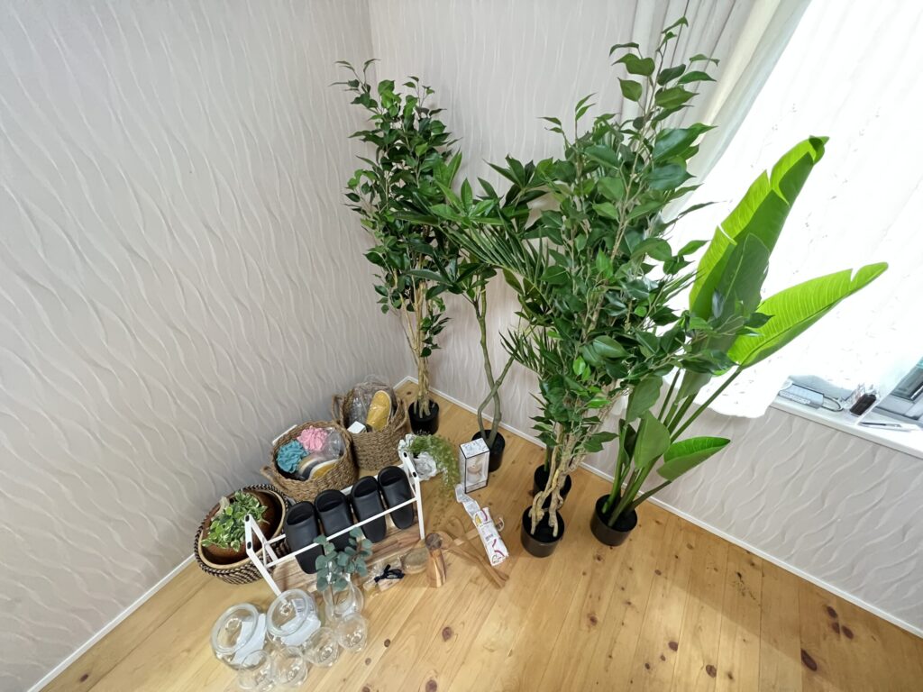 居室コーナーの室内装飾例。ウォールナット調アクセントウォールの前にジグザグ模様のバスケット入り観葉植物を置き、Bedroom（2）の設備仕様を記載したアクリルPOPスタンドを設置している