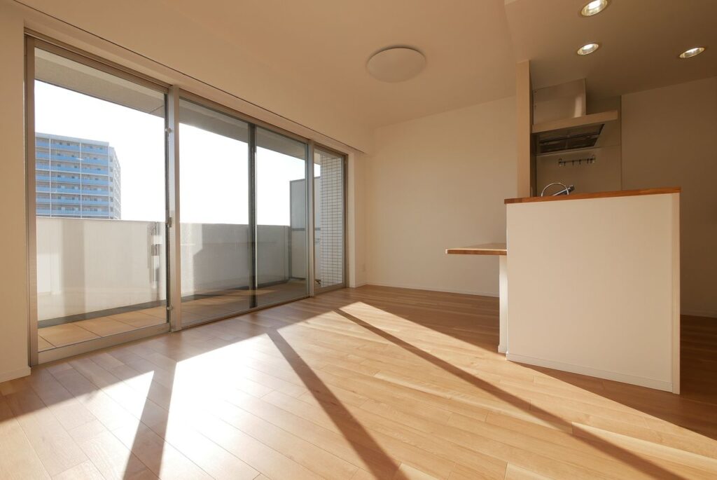 フローリング張り替え後のLDK。明るいナチュラルカラーの新しいフローリングが広がり、バルコニー側の大きな窓から日差しが差し込んでいる。右手に対面式キッチンのカウンターが見える