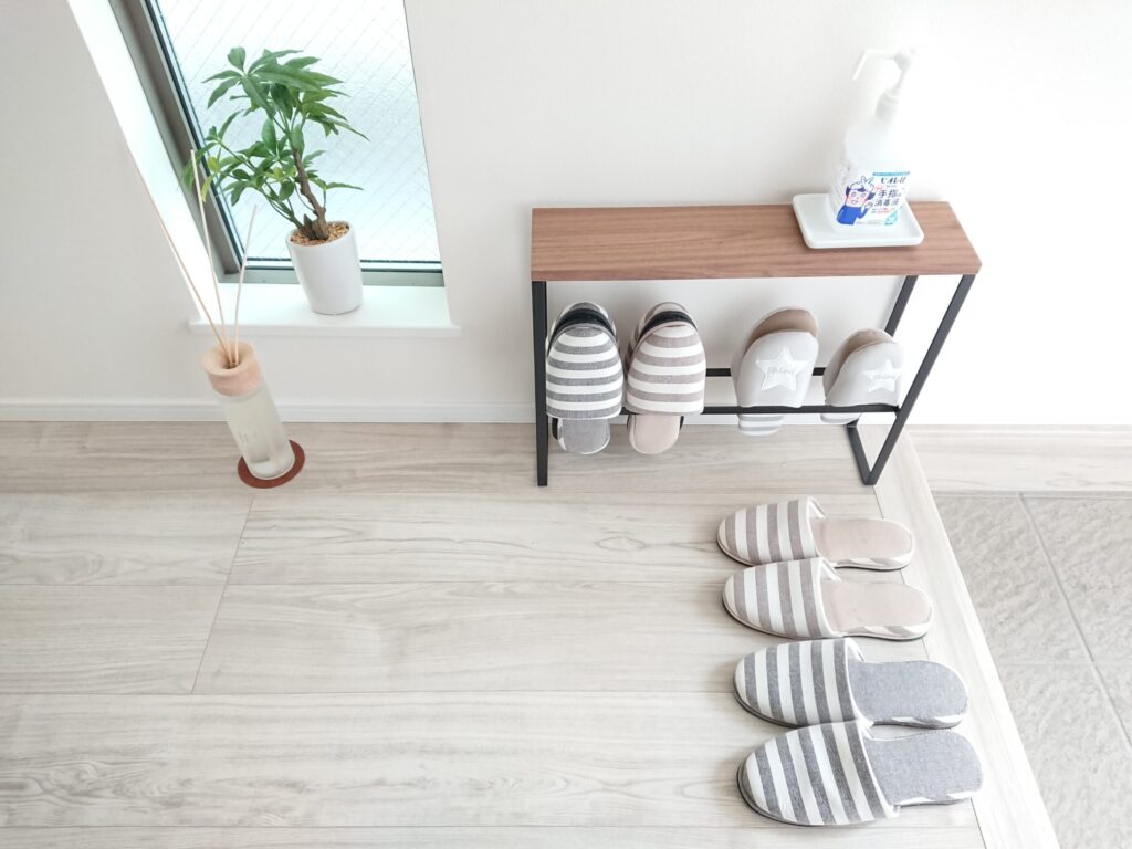 玄関の室内装飾例。ストライプ柄のスリッパを揃え、アイアン脚のシューズラック上に消毒液を置き、窓辺に観葉植物とリードディフューザーを飾っている